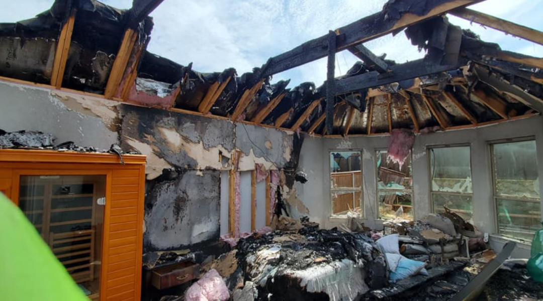 Interior of a fire-damaged room with charred walls, a collapsed roof, and debris scattered across the floor. Sunlight streams in through broken windows.